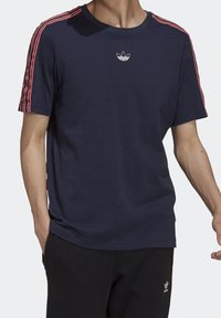 Mörkblå t-shirt med röda och vita ränder på axlarna, rund halsringning och litet broderat logotyp på bröstet; mjukt tyg, korta ärmar.