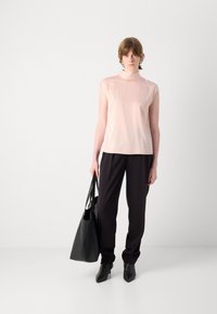 Top senza maniche di un delicato rosa pallido con colletto alto, abbinato a pantaloni neri su misura. La modella porta una grande borsa tote nera. Stivali neri a punta.