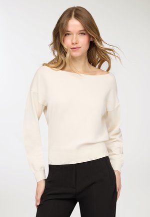 Maglione beige chiaro, a spalle scoperte, realizzato in tessuto morbido con maniche lunghe e fascia aderente in vita, abbinato a pantaloni neri.