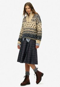 Dikke crème trui met marineblauwe geometrische patronen, gelaagd over een blauwe geruite blouse, gecombineerd met een donker geruite rok en bruine veterschoenen.