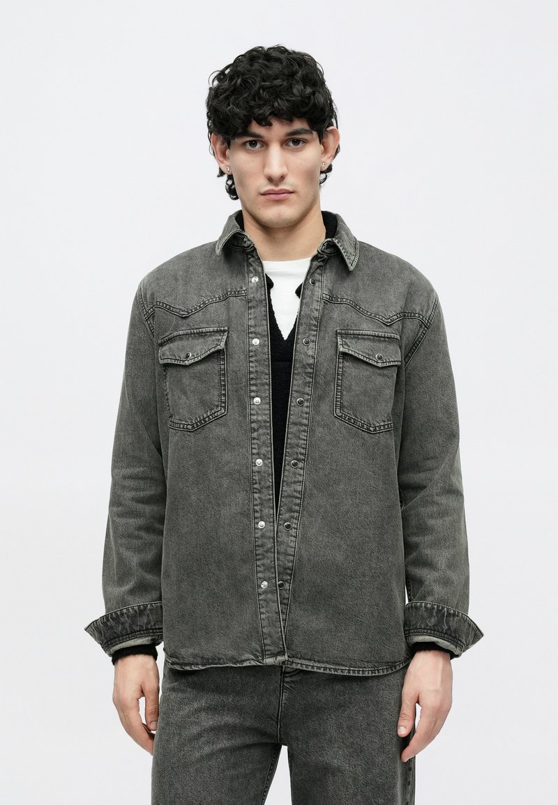 Young man with curly dark hair wearing a dark gray denim jacket and matching jeans over a black and white layered top.