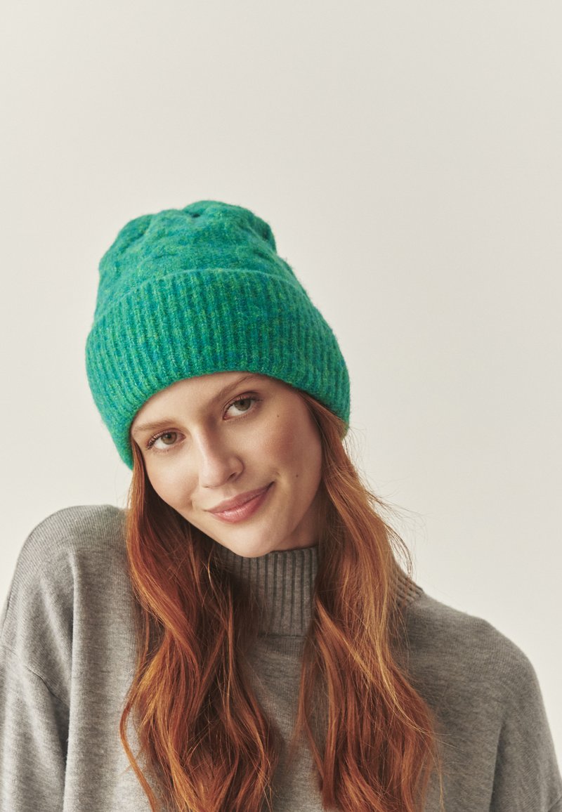 Grøn strikket hue lavet af tekstureret uld, med en ribbet kant og en let hængende top. Båret med en grå turtleneck trøje.