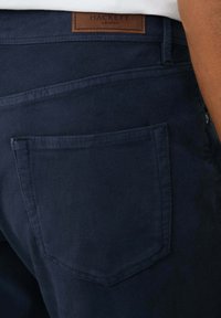 Marineblauwe broek van stof met een zachte textuur, met een achterzak en een bruine leren label met "HACKETT LONDON."