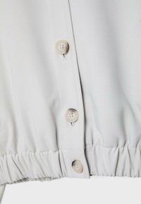 Gros plan sur l'ourlet d'une chemise blanche avec trois boutons beiges et une ceinture élastiquée.