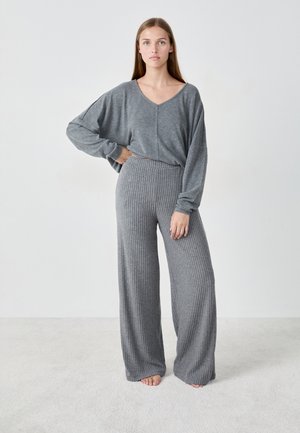Vrouw staat blootsvoets op een tapijt in een losse grijze V-hals trui en wijde ribgebreide broek tegen een effen witte achtergrond.