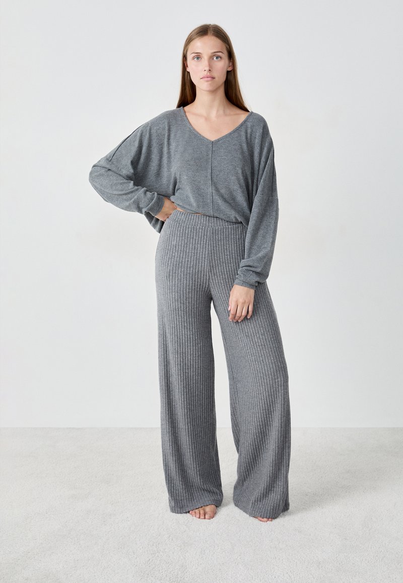 Kobieta stoi boso na dywanie, ubrana w luźny szary sweter z dekoltem w serek i szerokie, prążkowane spodnie, na tle gładkiej białej �ściany.