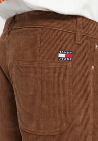 Pantalones de pana marrones con una textura acanalada, que cuentan con un parche de logo en blanco y rojo marino que dice "TOMMY JEANS" en el bolsillo trasero.