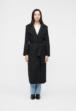 Nő, aki hosszú fekete, övvel ellátott ballonkabátot visel világoskék farmernadrág és fekete cipő fölött, egy egyszerű fehér háttér előtt áll.