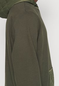 Sweat à capuche vert avec des détails en tissu matelassé sur l'épaule et la poche, montré de profil sur une personne.