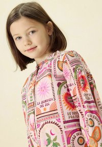 Blouse multicolore en rose, présentant des motifs botaniques et solaires. Fabriquée en tissu léger avec un col arrondi et une fermeture à boutons.