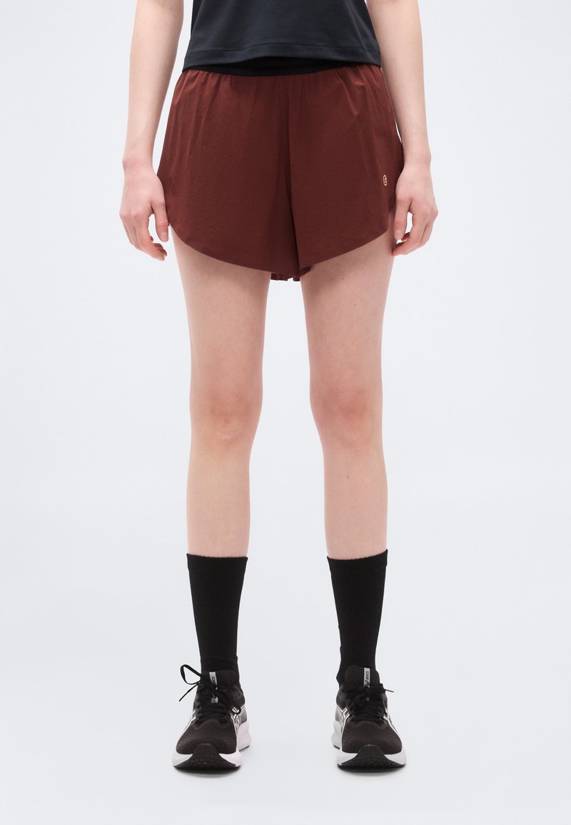 Άτομο που φοράει μαύρο αθλητικό σορτς μπορντό, μαύρες κάλτσες και μαύρα παπούτσια τρεξίματος, στέκεται μπροστά σε απλό φωτεινό φόντο.