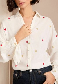 Camicia bianca con bottone, decorata con ricami a forma di cuore rossi e dorati, presenta maniche a sbuffo con polsini legati e una vestibilità rilassata, abbinata a jeans scuri.