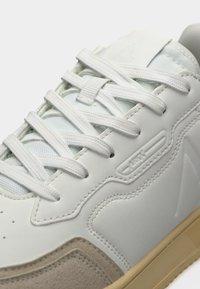 Λευκό αθλητικό παπούτσι με λευκά κορδόνια, μπεζ δερμάτινη μύτη, και φωτεινή καφέ σόλα, με λεπτομερή ραφές και ανάγλυφο λογότυπο στο πλευρικό πάνελ.