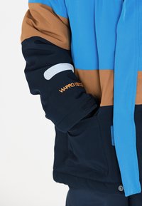 Färgglad jacka med blå, brun och marin paneler, strukturerad tyg, kardborre-muddar och logotypdetalj på ärmen i orange text.