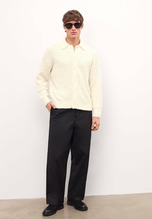 Cardigán de textura ligera en color crema claro con cierre de botones, combinado con unos pantalones negros holgados y botas negras, sobre un fondo blanco liso.