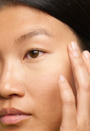 Gros plan d'un visage montrant une peau lisse et brun clair avec de légères taches de rousseur ; une main touche doucement la joue. La lumière naturelle met en valeur la texture de la peau.