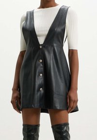 Vestido de cuero sintético negro con un escote en V profundo, diseño sin mangas y botones decorativos en la parte delantera. Parte superior texturizada de punto blanca debajo.