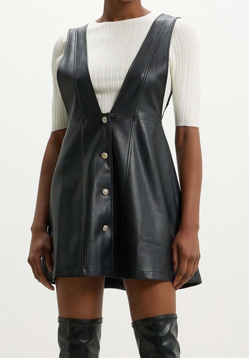 Vestido de cuero sintético negro con un escote en V profundo, diseño sin mangas y botones decorativos en la parte delantera. Parte superior texturizada de punto blanca debajo.