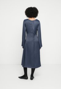 Rochie din satin bleumarin cu mâneci lungi și decolteu în V pe spate, prevăzută cu fermoar la spate și o siluetă fluidă. Purtați cu balerini negri și ciorapi.