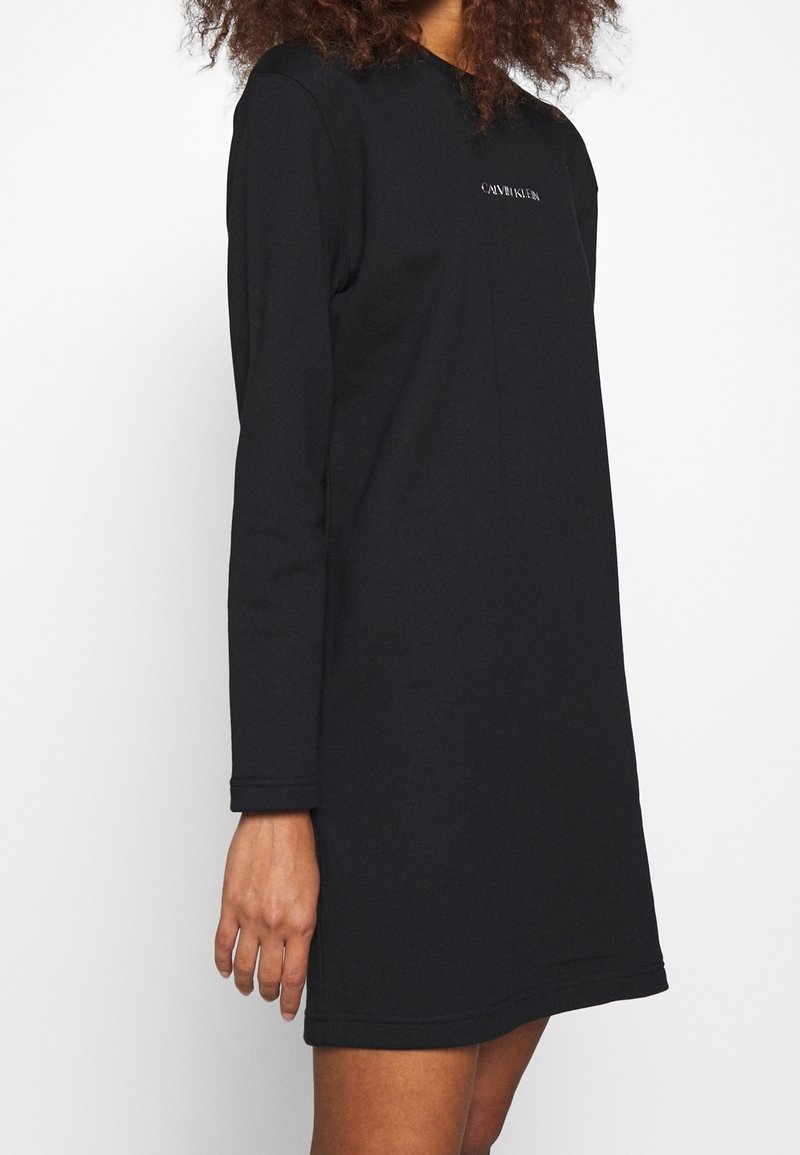 Vestido negro de manga larga hecho de tela suave. Presenta un escote redondo y un logo bordado en el pecho. Corte ajustado con una silueta recta.