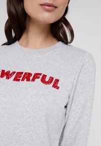 Sudadera gris de tela suave con cuello redondo. Presenta un texto rojo llamativo "PODEROSO" en una fuente texturizada y juguetona.
