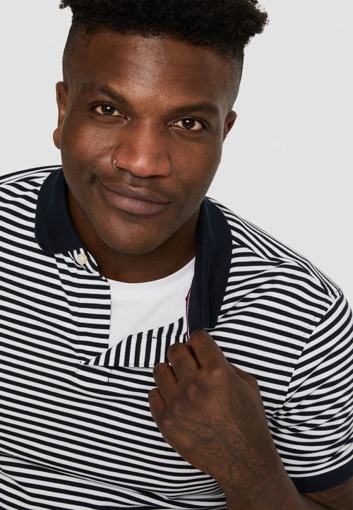 Polohemd mit marineblauem Kragen und Knopf, ausgestattet mit horizontalen Streifen in Marineblau und Weiß. Das Model hält den Kragen und zeigt ein weißes Unterhemd.