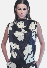 Top senza maniche nero con un motivo floreale bianco, con scollo drappeggiato e texture liscia. Il modello posa con le mani in tasca.