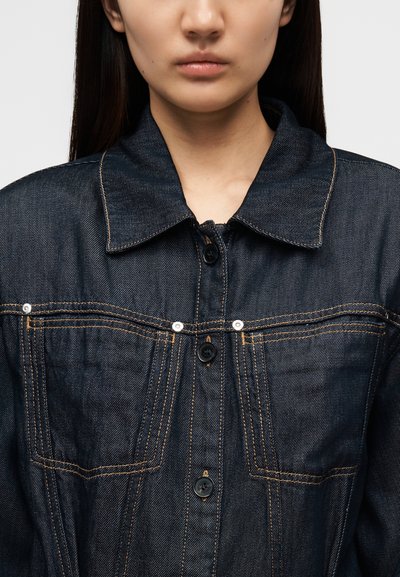 Σκούρο μπλε τζιν μπουφάν με γιακά, κλεισίματα με κουμπιά και εμπρόσθιες τσέπες. Διακρίνονται ραφές σε αντίθεση και υφή στο ύφασμα.