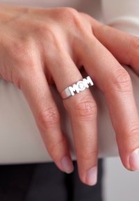 Silberring mit der Aufschrift "MOM", glatte Oberfläche, minimalistisches Design, an einem Finger mit heller Hautfarbe getragen.