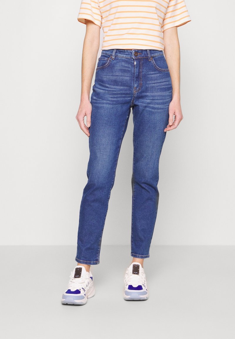 Personne portant un jean slim taille haute bleu, des baskets blanches et bleues, et un t-shirt à manches courtes rayé jaune et blanc.