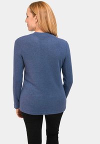 Blaue Langarmbluse mit Rundhalsausschnitt, aus weichem, strukturiertem Stoff. Der Rücken ist glatt und etwas länger, kombiniert mit schwarzen Hosen.