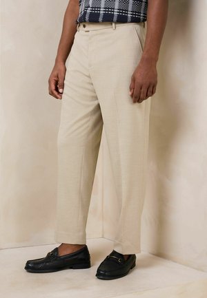 Mand iført beige skræddersyede bukser, sorte læder loafers og en navy mønstret skjorte, der står op ad en lys væg.