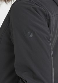 Musta pehmeäkuorinen takki, jossa on sileä pinta, tyylikäs muotoilu ja sävy sävyyn -logo hihassa. Kevyesti topattu lämmön takaamiseksi.