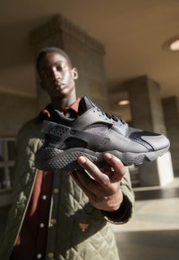 Zapatilla deportiva negra con suela texturizada, paneles de malla y material sintético suave. Cuenta con un collar acolchado y un sistema de cordones.