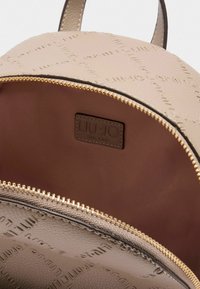 Interno di uno zaino in pelle beige con un motivo a logo in rilievo e dettagli in oro; fodera marrone con un'etichetta del marchio.