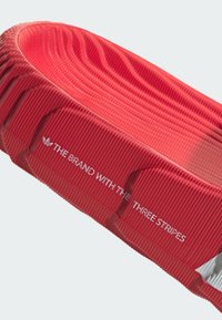 Red textured sandal with ridged surface. Features logo text “THE BRAND WITH THE THREE STRIPES.” Smooth interior lining.