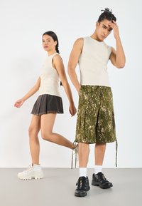 Beige ribots bez piedurknēm, melna džinsa plisētā svārki un zaļi kamuflāžas kravas šorti; savienoti ar baltām un melnām masīvajām sporta kurpēm.