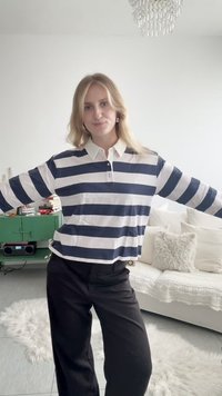 Jeune femme aux cheveux blonds portant une chemise rayée noire et blanche et un pantalon noir, debout les bras étendus dans un salon lumineux.
