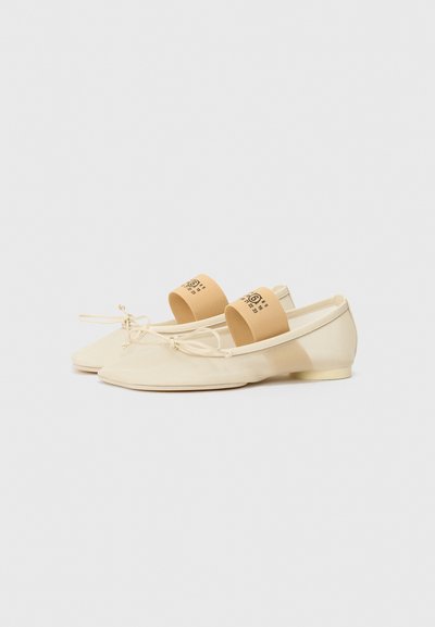 Light beige ballet flats with a rounded toe, elastic straps, a soft material finish, and a small heel. Accented with a logo on the strap.