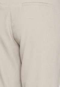 Gros plan sur un pantalon en tissu beige montrant les détails des coutures et une seule poche arrière du côté droit.