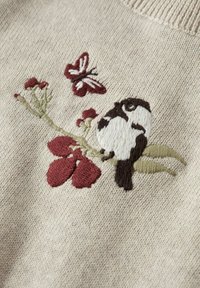 Moineau brodé perché sur une branche avec des fleurs rouges et un papillon rouge sur un fond de tissu tricoté beige.