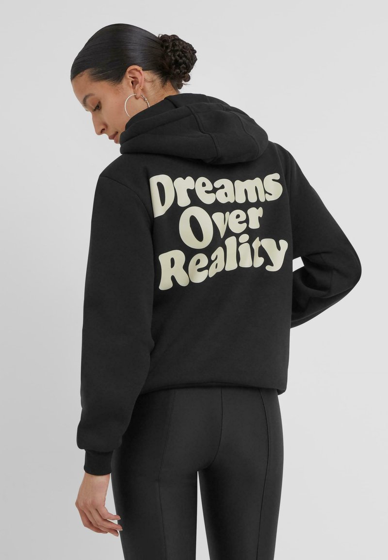 Zwarte hoodie met een crèmekleurige grafische tekst op de achterkant die luidt "Dromen boven de werkelijkheid." Zacht materiaal, ontspannen pasvorm.