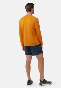 Langärmliges oranges Hemd, marineblauen gemusterte Shorts und schwarze Schuhe. Das Hemd hat eine glatte Textur; die Shorts weisen ein lebhaftes abstraktes Design auf.