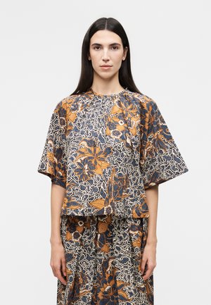 Γυναίκα με μακριά σκούρα μαλλιά φοράει χαλαρή κορυφή και φούστα με ταιριαστό μοτίβο με περίπλοκα σχέδια σε πορτοκαλί, ναυτικό μπλε και λευκές αποχρώσεις λουλουδιών και φύλλων.