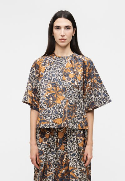 Woman with long dark hair wears matching loose floral top and skirt featuring orange, navy, and white intricate flower and leaf patterns.