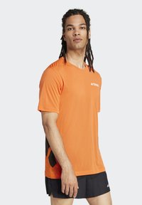 Camiseta deportiva naranja de manga corta, con cuello redondo y paneles laterales negros texturizados que presentan un patrón geométrico. Logotipo en el pecho.