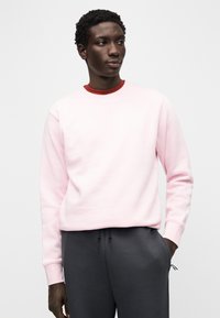 Sweatshirt rosa claro com gola redonda, punhos canelados e logótipo branco. Combinado com calças de fato de treino cinzentas escuras com detalhe de cordão.