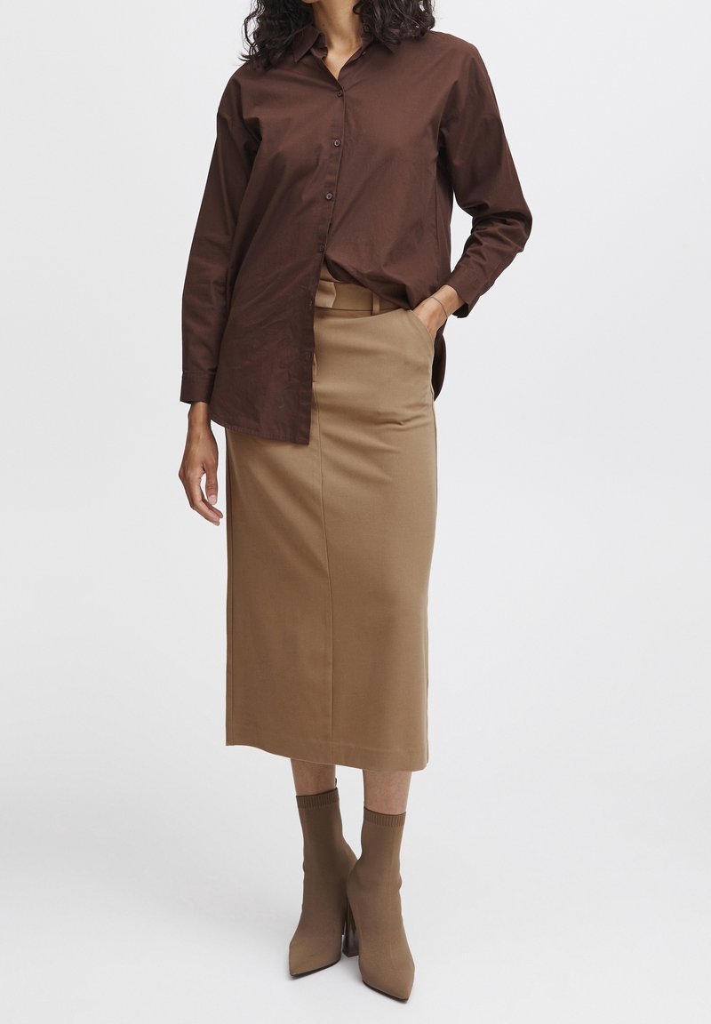Femme portant une chemise marron foncé boutonnée rentrée dans une jupe midi beige avec des poches latérales et des bottines marron, debout avec une main dans une poche.