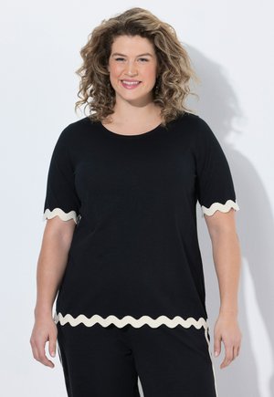 Donna sorridente con capelli ricci, indossa un top nero e pantaloni con bordi ondulati bianchi su maniche e orlo, in piedi su uno sfondo bianco.