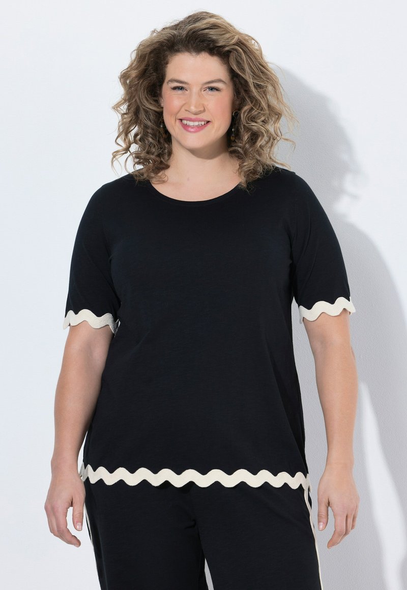 Donna sorridente con capelli ricci, indossa un top nero e pantaloni con bordi ondulati bianchi su maniche e orlo, in piedi su uno sfondo bianco.